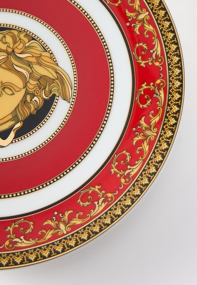 Πορσελάνινο πιάτο κόκκινου και λευκού χρώματος με κεντρικό σχέδιο χρυσού προσώπου, περιτριγυρισμένο από περίτεχνα χρυσά και μαύρα διακοσμητικά κατά μήκος της στεφάνης.