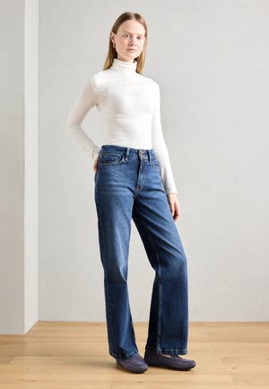 Jeans skinny taille haute en denim bleu foncé. Accompagnés d'un col roulé blanc ajusté et de chaussures glissantes grises. Texture lisse et coupe classique.