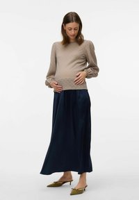 Maglia premaman in tessuto beige testurizzato con maniche lunghe, abbinata a una gonna maxi fluida blu navy e mule verdi oliva con punta affusolata.