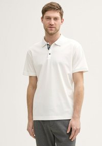 Weißes Polohemd mit kurzen Ärmeln und schwarzem Etikett am Kragen, mit einer Knopfleiste mit zwei Knöpfen und glattem Material, kombiniert mit grauen Hosen.
