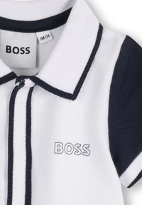 T-shirt polo blanc avec des accents navy, présentant un col, une patte de boutonnage et un logo "BOSS" brodé sur la poitrine gauche. Tissu texturé.