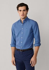 Junger Mann, der ein blaues Hemd mit hochgekrempelten Ärmeln, dunkelblaue Hosen und einen schwarzen Gürtel trägt, steht vor einem einfarbigen Hintergrund.