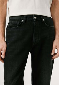 Vaqueros de denim negros con un ajuste clásico, que presentan un diseño de cinco bolsillos, cierre frontal con botón y un sutil desgaste en la tela.
