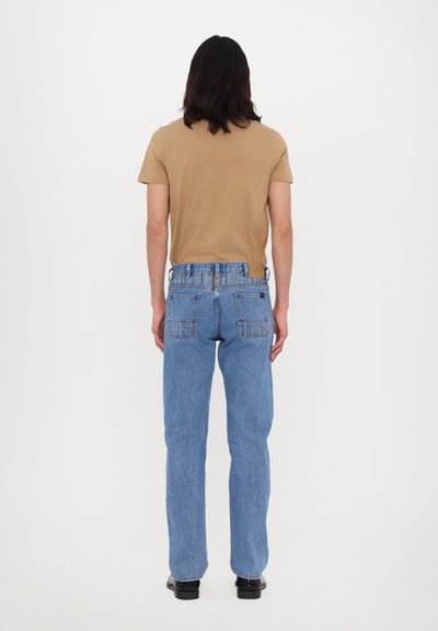 Light brown short-sleeve t-shirt, blue denim jeans with straight-leg fit, visible back pockets, and smooth texture. Black footwear completes the look.