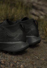 Paire de chaussures de trail noires et mouillées reposant sur un chemin de gravier avec de l'herbe verte floue en arrière-plan.