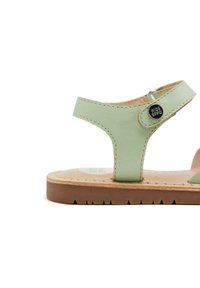 Sandal in pelle verde chiaro con design a T, suola in materiale brown texturizzato e un bottone metallico vicino alla cinghia alla caviglia.