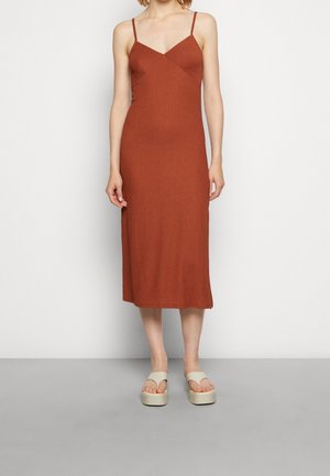 Robe midi côtelée couleur rouille avec fines bretelles, décolleté en V, coupe ajustée et fentes latérales, portée avec des sandales neutres.