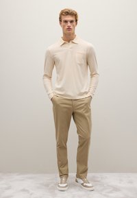 Camisa polo de manga larga en beige claro con un bolsillo en el pecho, combinada con pantalones beige a medida y zapatillas de color claro con acentos texturizados.