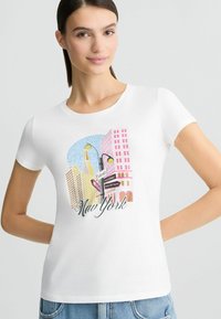 T-shirt blanc à manches courtes présentant un graphique coloré du skyline de New York, avec des bâtiments dans des teintes pastel et des détails de panneaux de signalisation.