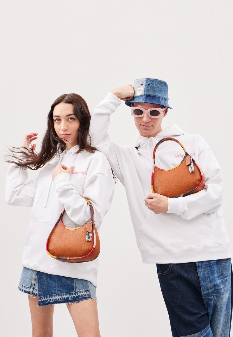 Väriltään tan, jossa moniväriset reunakoristeet, teksturoitu pinta ja koristeellinen tunniste. Kaksi mallia pukeutuneena valkoisiin huppareihin ja farkkushortseihin.