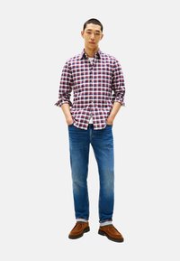 Camisa de cuadros roja y azul con botones, jeans azul claro, zapatos de ante marrón. Modelo de pie con las manos en los bolsillos sobre un fondo blanco.