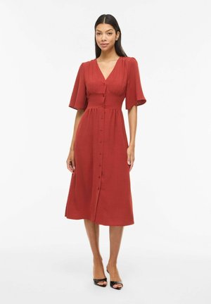 Femme debout portant une robe midi rouge rouille avec des manches courtes bouffantes et des boutons à l'avant, associée à des talons ouverts noirs sur fond blanc.
