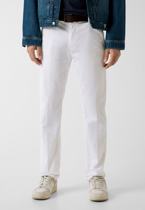 Homme portant un pantalon blanc, des baskets beiges, une ceinture noire, une veste en jean foncée et une chemise noire, debout devant un fond uni.