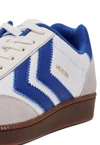 Białe sneakersy z niebieskimi akcentami z materiału syntetycznego i zamszowego, wzorem w zygzaki, okrągłym noskiem, płaskimi sznurowadłami i brązową gumową podeszwą.