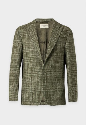 Mann steht und trägt einen grünen strukturierten Blazer, weißes Hemd, braune Hose und dunkelbraune Lederschuhe vor weißem Hintergrund.