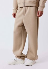 Beige, loszittende trainingsbroek met elastische boorden, gedragen met witte sneakers met zwarte details, getoond op een staande persoon.