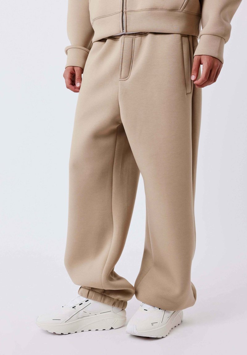 Beige, loszittende trainingsbroek met elastische boorden, gedragen met witte sneakers met zwarte details, getoond op een staande persoon.