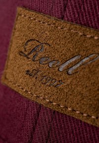 Tela marrón con un parche texturizado en marrón que presenta un grabado en relieve de "Reell" y "est. 1997" en color plata. Bordes cosidos para un acabado detallado.