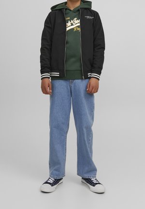 Giacca nera con zip e polsini a strisce bianche sopra un hoodie verde. Jeans in denim azzurro chiaro e sneakers nere con suola bianca.