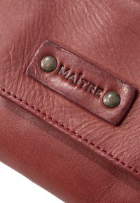 Rote Lederbörse mit strukturierten Oberflächen, ausgestattet mit einem Logo-Patch, Metallnieten und kontrastierenden Nähten entlang des Randes.