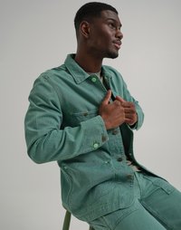 Veste rayée verte en tissu texturé, dotée de boutons, de poches poitrine et d'un col. Le modèle pose avec les mains près de la veste.