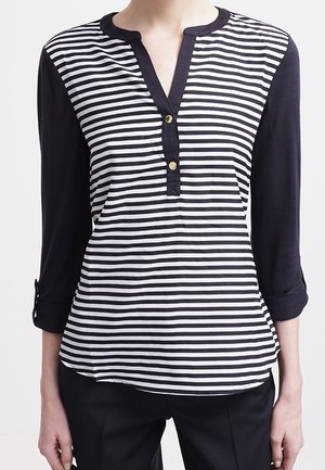 Vrouw draagt een zwart-wit gestreept shirt met horizontale strepen, zwarte mouwen en twee gouden knopen bij de halslijn.