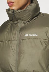 Gros plan sur une personne portant une doudoune Columbia vert olive avec un col montant et une fermeture éclair avant visible.