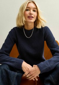 Maglione a maglia blu navy con scollo rotondo e maniche lunghe; tessuto testurizzato; il modello è seduto su una poltrona in pelle marrone, indossando gioielli semplici.