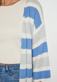 Gestrickte Strickjacke mit abwechselnden blauen, grauen und weißen Streifen. Weiche Textur, offene Vorderseite und lässige Passform, kombiniert mit einem figurbetonten beigen Top.