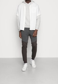 Grijze hoodie gedragen onder een kortmazige witte overhemd, gecombineerd met donkergrijze en zwarte kleuren-blok joggers, en witte sneakers.