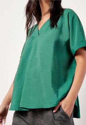 Vrouw draagt een losse groene V-hals top met korte mouwen en een gestructureerde stof, gecombineerd met grijze geruite broek, hand in zak.