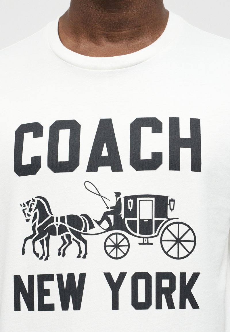 Chemise blanche avec texte noir "COACH NEW YORK" et graphique d'une calèche tirée par des chevaux avec un cocher tenant les rênes et un fouet.