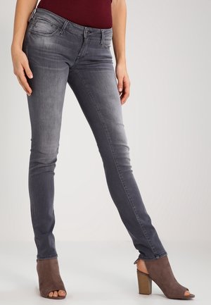 Žena v opraných šedých úzkých džínách, bordovém topu a hnědých otevřených kotníkových botách na podpatku stojí před jednobarevným pozadím.