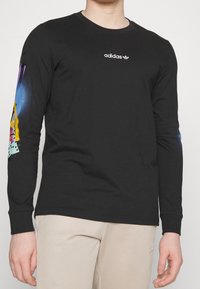 Mężczyzna w czarnej koszulce z długim rękawem Adidas z kolorowymi grafikami na prawym rękawie i beżowych spodniach.