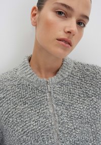 Un giovane con la pelle chiara indossa un maglione grigio zip-up testurizzato, rivolto leggermente verso l'alto su uno sfondo chiaro e semplice.