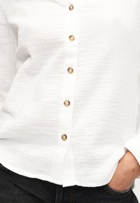 Chemise blanche texturée à manches longues avec des boutons marron portée par une personne avec une main dans la poche d'un jean foncé, cadrée à la taille.