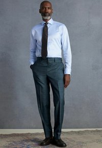 Lyseblå skjorte med lange ærmer, mønstret mørk slips og mørkegrønternede bukser. Sorte sko fuldender outfittet.
