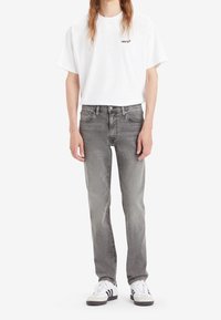 Jeans in denim grigio con vestibilità classica, abbinati a una maglietta a maniche corte bianca con un piccolo logo nero. Sneakers bianche con dettagli neri.