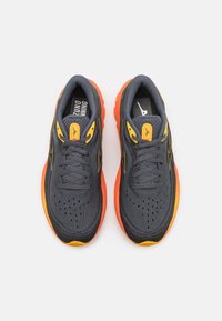 Scarpe da ginnastica grigie con suole arancioni, dotate di una tomaia in rete testurizzata, design con lacci e distintive accenti gialli sulla linguetta e sui lati.