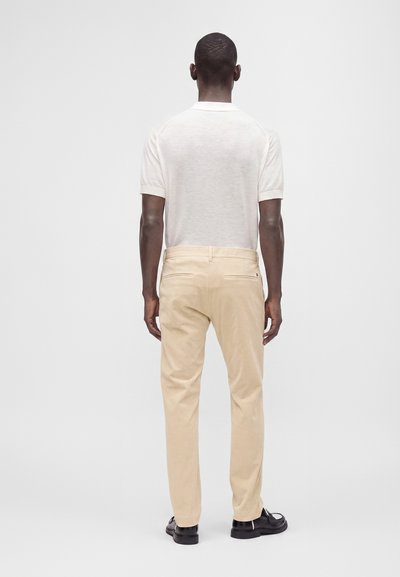 Homme portant une chemise blanche à manches courtes, un pantalon beige et des chaussures noires, debout de dos devant un fond clair uni.