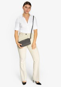 Borsa a tracolla nera realizzata in materiale testurizzato, forma rettangolare, con una lunga tracolla, appoggiata su una camicia bianca e pantaloni beige a campana.