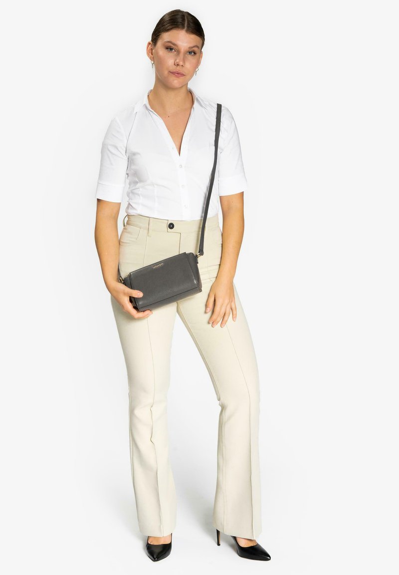 Borsa a tracolla nera realizzata in materiale testurizzato, forma rettangolare, con una lunga tracolla, appoggiata su una camicia bianca e pantaloni beige a campana.
