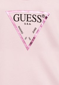 Rosa t-skjorte med et stort, trekantet logo i blank rosa, der det står "GUESS", "WASHED" og "JEANS" i svart tekst. Myk stoffkvalitet.