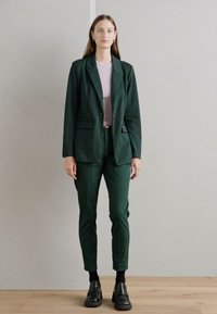 Mörkgrön skuren kavaj och matchande byxor, bärs över en ljusrosa topp. Outfiten har eleganta linjer och subtila fickdesigns.