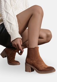Bottines en daim marron avec une semelle texturée, un talon carré et des accents de couture verticale. Le design comprend une languette pour un enfilage facile.