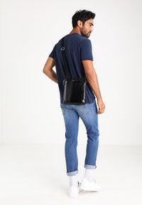 Bolso cruzado de cuero negro con correa ajustable, que cuenta con un bolsillo con cremallera en la parte delantera y detalles en azul. Modelo lleva puestos jeans azules y zapatos.