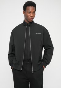 Μαύρο μπουφάν bomber με φερμουάρ μπροστά, πλεκτό γιακά και μανσέτες. Διαθέτει διακριτική λευκή επωνυμία και τσέπες στο πλάι.