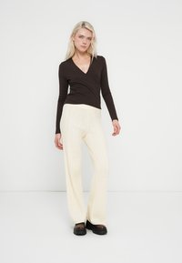 Top marrone a maniche lunghe con incrocio, abbinato a pantaloni ampi a coste color crema. L'outfit presenta una silhouette aderente e un tessuto strutturato, completato da scarpe nere chunky.