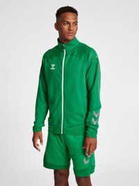 Grøn atletisk jakke med lynlås foran, høj krave og mesh-struktur; parret med matchende shorts, har reflekterende accenter og logo.