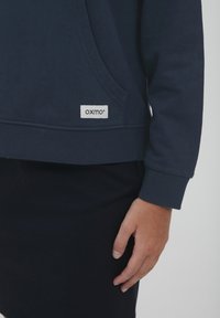 Tamsiai mėlynas megztinis su kišene, su įtempimais ant rankovių ir baltu etiketu su "oxmo" logotipu netoli apačios. Lygi tekstūra.